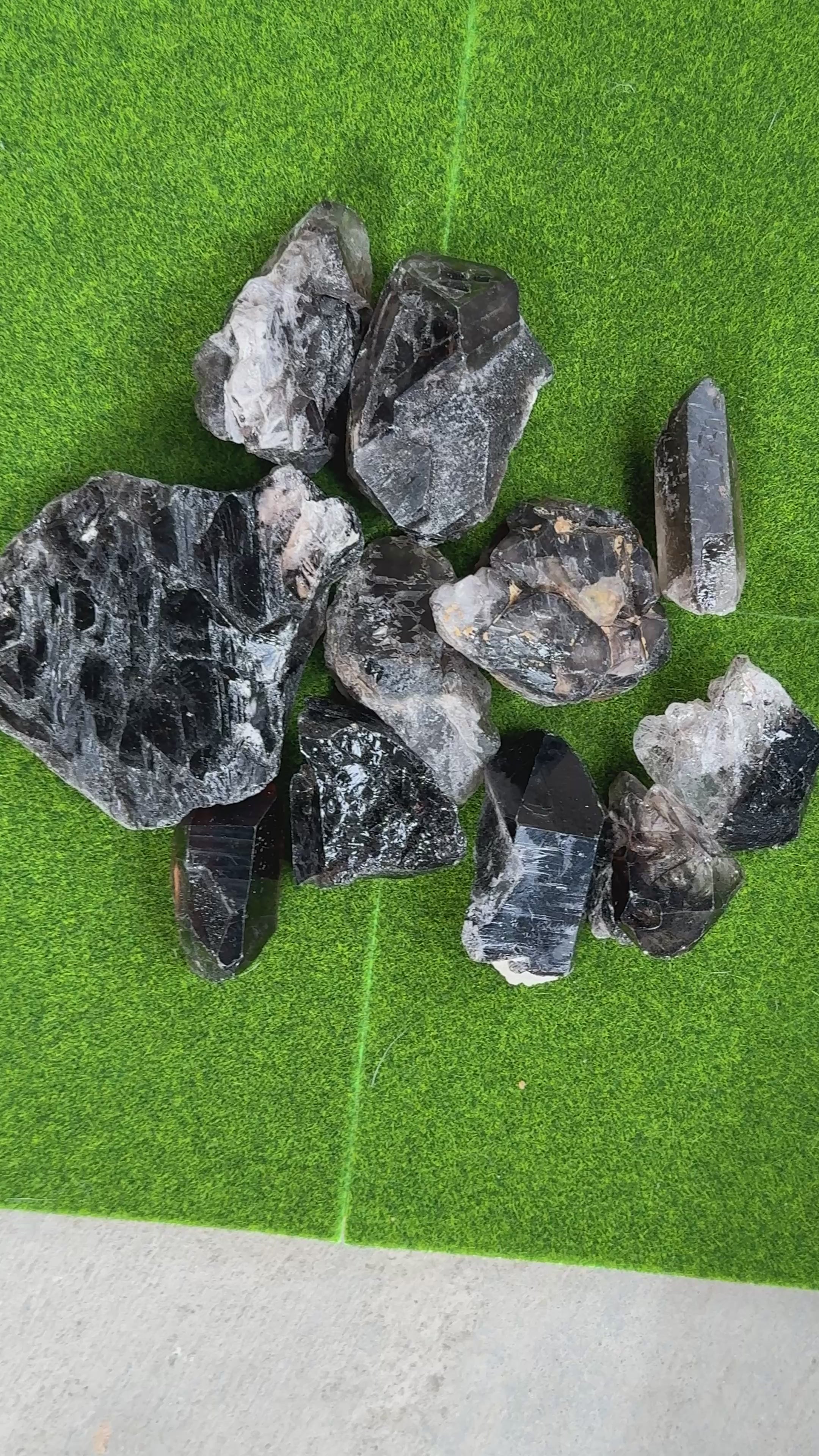 A collection of raw, smoky quartz crystals in various shapes and sizes. The crystals range in color from dark brown to translucent gray, with visible natural textures and facets. The crystals are arranged on a green surface.