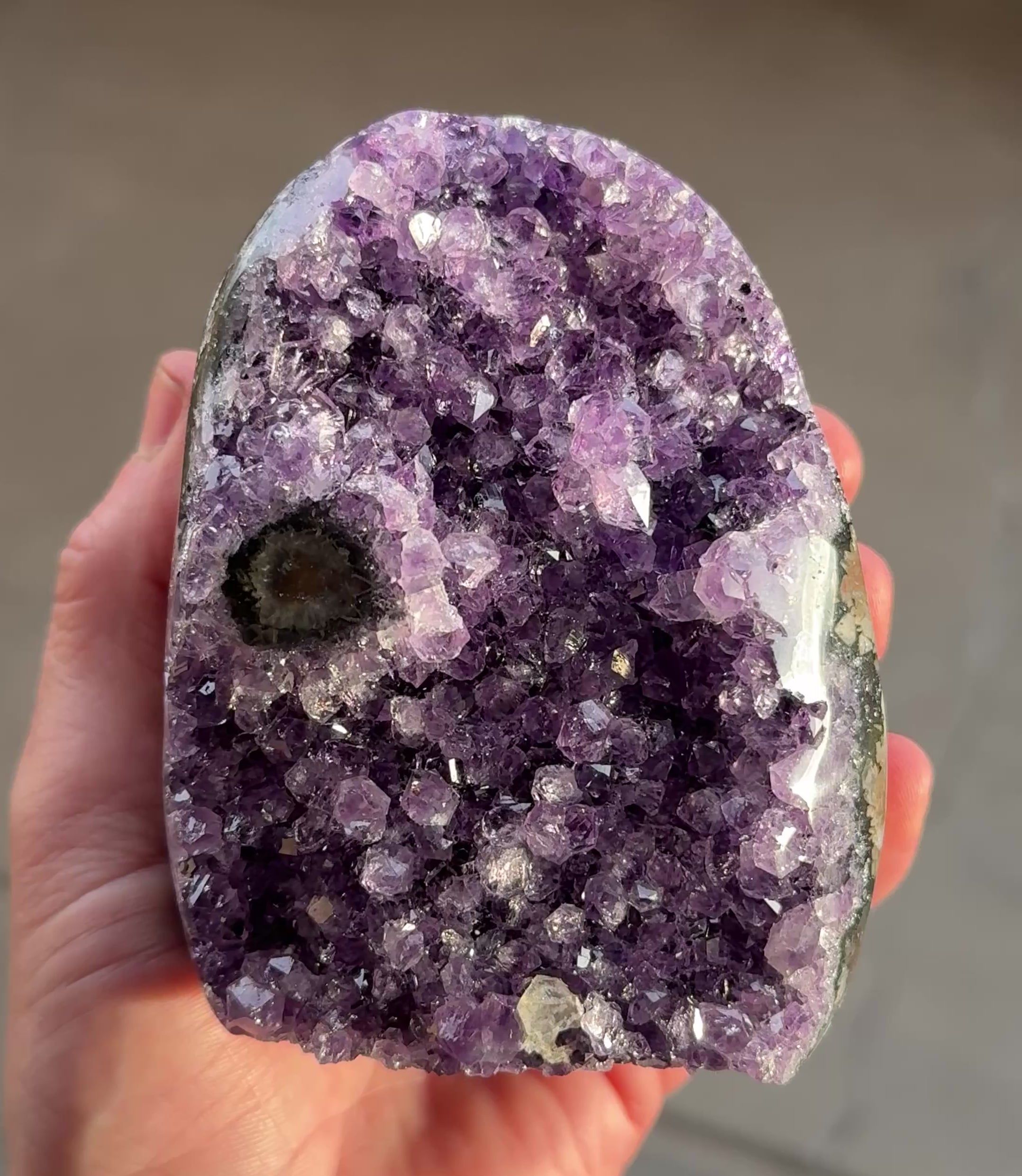 A polished amethyst geode with a deep purple hue. The crystal formation is visible, with numerous small, sparkling points. A dark, circular inclusion is present near the base, adding contrast to the overall color. The geode is a natural, decorative item.